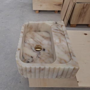 May include: A rectangular marble sink featuring a cream and beige veined pattern with a fluted exterior design. The basin includes a polished gold-toned circular drain cover centered at the bottom, showcasing natural stone variations throughout the surface.
