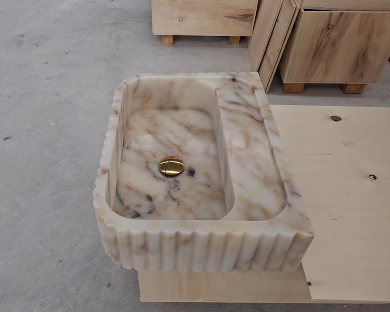 May include: A rectangular marble sink featuring a cream and beige veined pattern with a fluted exterior design. The basin includes a polished gold-toned circular drain cover centered at the bottom, showcasing natural stone variations throughout the surface.