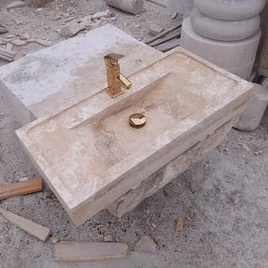 May include: A rectangular beige stone sink featuring a polished gold-tone faucet and a matching gold-tone drain cover. The basin has a smooth, flat rim and a recessed center, showcasing the natural porous texture and light tan color of the stone.