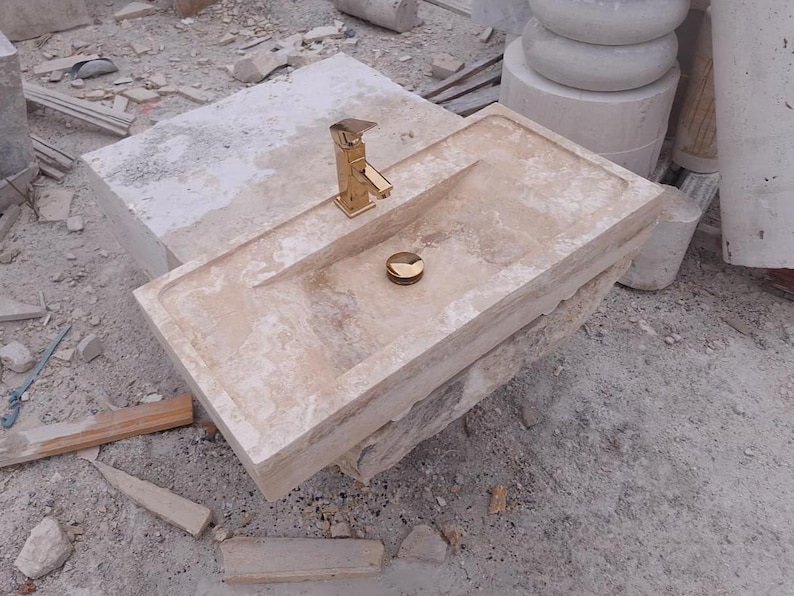 May include: A rectangular beige stone sink featuring a polished gold-tone faucet and a matching gold-tone drain cover. The basin has a smooth, flat rim and a recessed center, showcasing the natural porous texture and light tan color of the stone.