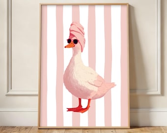May include: A framed art print featuring a whimsical illustration of a white duck wearing sunglasses and a pink towel on its head. The duck stands against a pink and white striped background. The artwork is displayed on a wooden floor.
