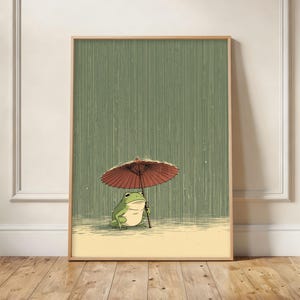 以下が含まれることがあります： 雨の中で赤い傘の下にいる緑のカエルを描いた額入りのアートプリント。カエルは漫画のスタイルで描かれており、穏やかな表情をしています。背景は緑とベージュの色合いの垂直ストライプパターンです。