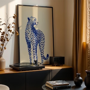 May include: A framed vertical art print featuring a blue cheetah illustration with dark blue spots on a light beige background. The print is displayed in a thin black frame, positioned behind a black record player on a wooden cabinet surface.