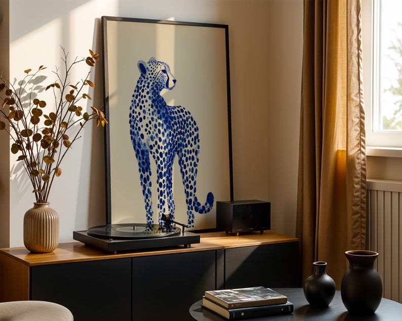 May include: A framed vertical art print featuring a blue cheetah illustration with dark blue spots on a light beige background. The print is displayed in a thin black frame, positioned behind a black record player on a wooden cabinet surface.