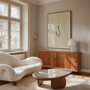 May include: A framed wall art print featuring a black fishing hook hanging from a thin chain against a cream background. The artwork is displayed above a wooden sideboard with sliding doors, a white table lamp, a curved white sofa, and a coffee table.