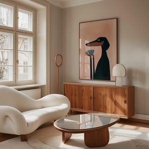 May include: A framed wall art print featuring a stylized black dog with long ears holding a small blue cup, displayed above a wooden sideboard with sliding doors, a white table lamp, a curved white textured sofa, and an oval glass coffee table.