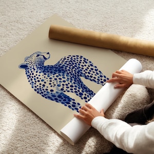 May include: A large square art print featuring a stylized blue cheetah illustration with distinct dark blue spots on a solid beige background. The print is shown partially rolled, with a separate brown cardboard mailing tube lying nearby.