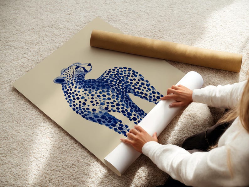 May include: A large square art print featuring a stylized blue cheetah illustration with distinct dark blue spots on a solid beige background. The print is shown partially rolled, with a separate brown cardboard mailing tube lying nearby.