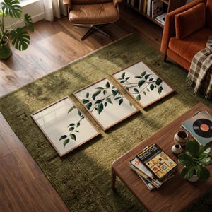 May include: A set of three rectangular wall art prints in thin wooden frames, each featuring a botanical illustration of green leaves on a light cream background, arranged in a row on a textured green rug in a living room setting.