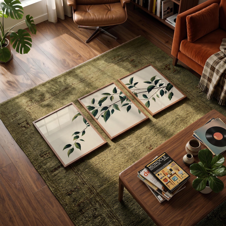 May include: A set of three rectangular wall art prints in thin wooden frames, each featuring a botanical illustration of green leaves on a light cream background, arranged in a row on a textured green rug in a living room setting.
