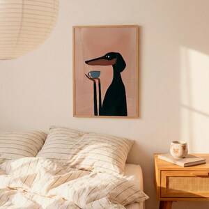 May include: A framed wall art print featuring a stylized black and tan dog with long limbs holding a small blue cup. The dog is depicted against a solid light pink background. The artwork is enclosed in a simple light-colored wooden frame.
