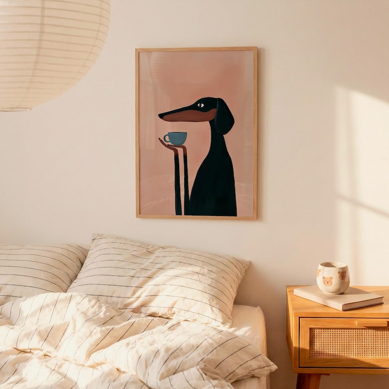 May include: A framed wall art print featuring a stylized black and tan dog with long limbs holding a small blue cup. The dog is depicted against a solid light pink background. The artwork is enclosed in a simple light-colored wooden frame.