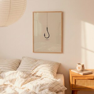 May include: A framed wall art print featuring a minimalist black line drawing of a single fishing hook hanging from a vertical line centered on a solid light beige background, enclosed in a simple light-colored wooden frame.