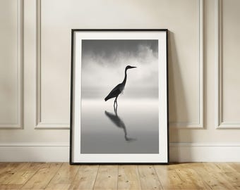 May include: A black and white framed wall art print featuring a silhouette of a heron standing in shallow water. The bird is captured in profile, with its reflection visible on the water surface against a soft, cloudy, and misty background.