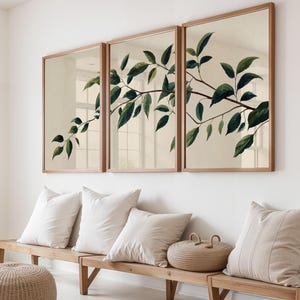 May include: A triptych wall art set featuring a continuous botanical illustration of a leafy branch with dark green leaves on a light beige background, framed in light wood, displayed above a wooden bench with several beige throw pillows.