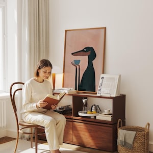 May include: A framed wall art print featuring a stylized illustration of a black dog with long ears and a snout, holding a small light blue cup in its hands against a solid dusty pink background, displayed in a thin wooden frame on a shelf.