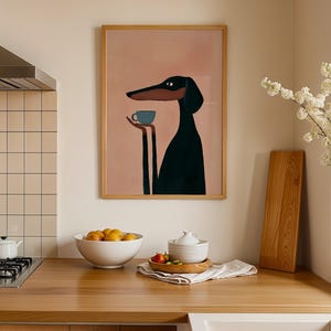 May include: A framed wall art print featuring a stylized illustration of a black dog with a long snout holding a small blue cup. The dog is set against a solid, muted pink background. The artwork is enclosed in a simple, light-colored wooden frame.