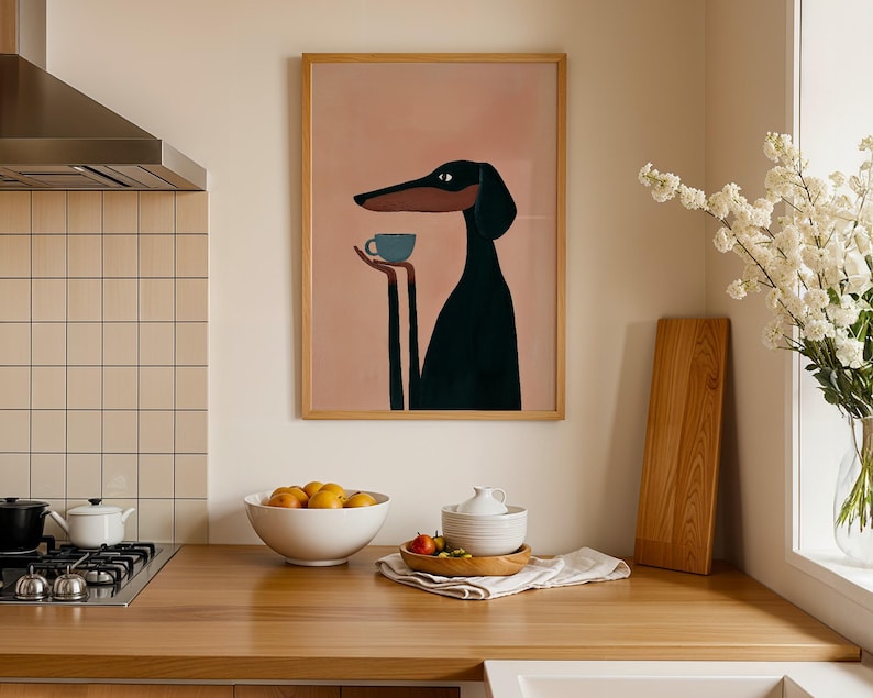 May include: A framed wall art print featuring a stylized illustration of a black dog with a long snout holding a small blue cup. The dog is set against a solid, muted pink background. The artwork is enclosed in a simple, light-colored wooden frame.