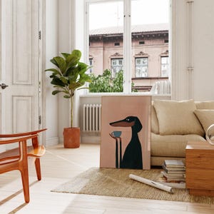 May include: A rectangular framed wall art print featuring a stylized illustration of a black dog with long ears and a long snout, holding a small light blue cup in its paws against a solid light pink background.
