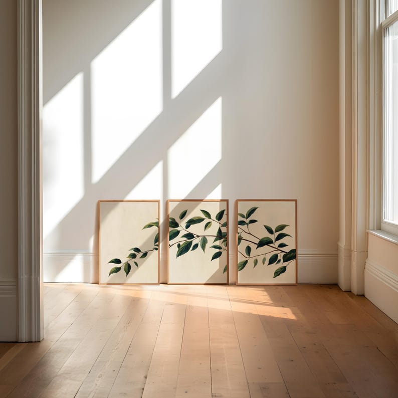 May include: A set of three rectangular wall art prints in light wood frames, each featuring a botanical illustration of a dark green leafy branch on a cream-colored background, arranged side by side on a wooden floor against a white wall.