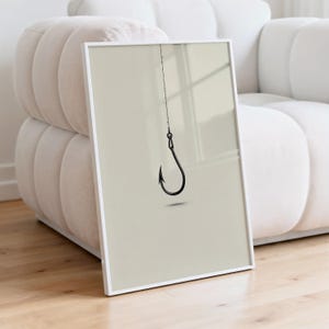 May include: A framed wall art print featuring a minimalist illustration of a single black fishing hook suspended by a thin line against a solid, light beige background. The artwork is encased in a simple, thin white frame.