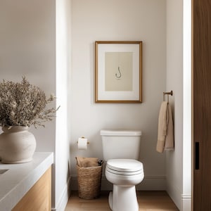 May include: A bathroom interior featuring a white toilet, a framed print of a fishing hook on the wall, a woven basket containing a tan cloth, a beige towel on a brass rack, and a large ceramic vase filled with dried foliage on a vanity.