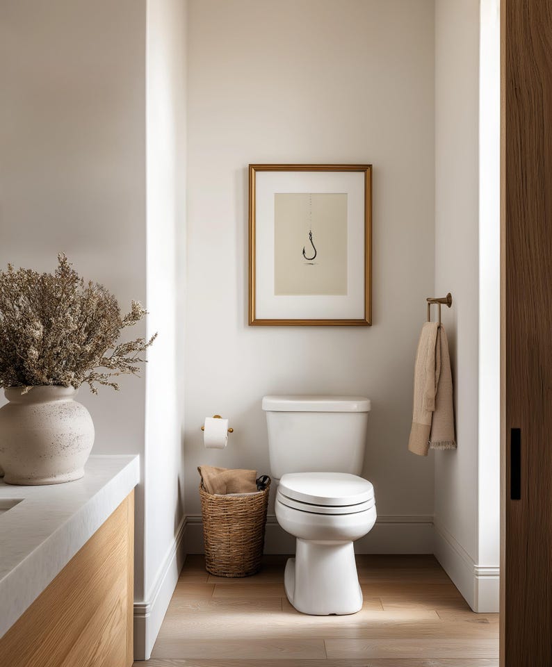 May include: A bathroom interior featuring a white toilet, a framed print of a fishing hook on the wall, a woven basket containing a tan cloth, a beige towel on a brass rack, and a large ceramic vase filled with dried foliage on a vanity.
