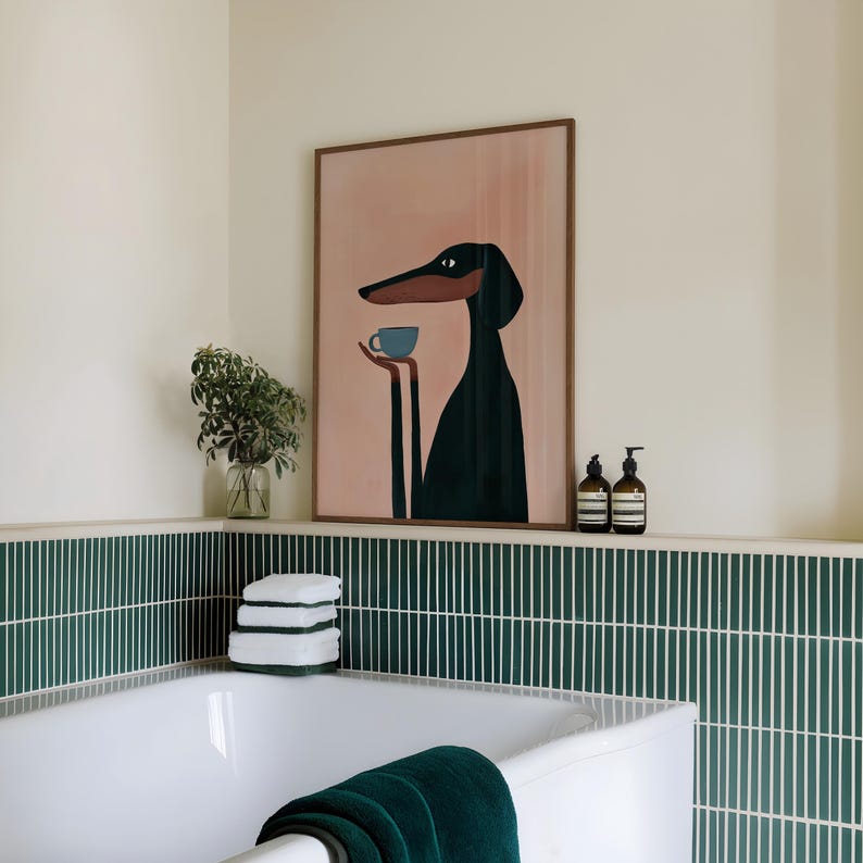 May include: A framed wall art print featuring a stylized black dog with long ears and a snout, holding a small light blue cup in its paws against a soft peach-colored background. The artwork is displayed in a thin wooden frame on a ledge.