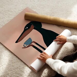 May include: A square art print featuring a stylized black and brown dog with long ears holding a small blue teacup in its paws. The background is a solid muted peach color. The print is partially rolled and rests next to a brown cardboard tube.