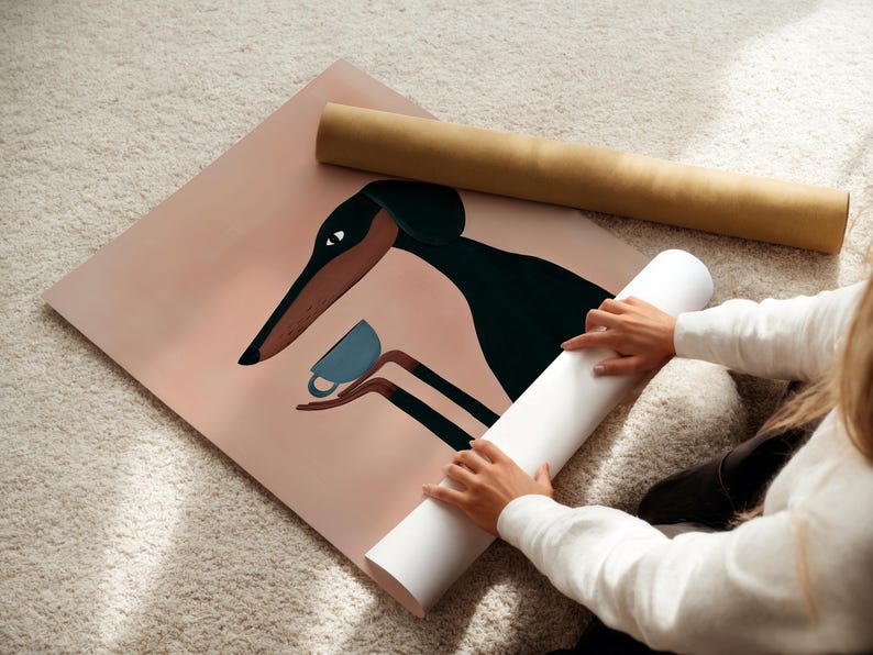 May include: A square art print featuring a stylized black and brown dog with long ears holding a small blue teacup in its paws. The background is a solid muted peach color. The print is partially rolled and rests next to a brown cardboard tube.