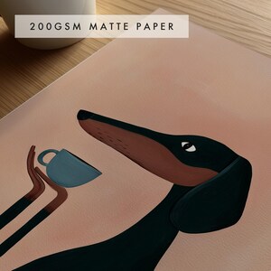 May include: A print on 200gsm matte paper featuring a stylized illustration of a black and brown dachshund dog looking toward a small blue cup held by two brown arms extending from the left side against a solid light peach background.