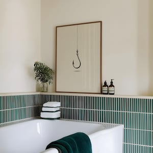 May include: A framed wall art print featuring a minimalist black fishing hook illustration suspended by a thin line against a light beige background, displayed in a bathroom with dark green vertical tile walls and a white bathtub.