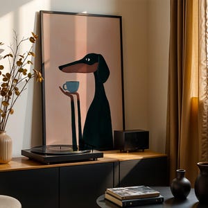 May include: A framed wall art print featuring a stylized illustration of a long-necked black dog with a tan muzzle holding a small light blue coffee cup. The background of the artwork is a solid soft peach color, enclosed in a thin black frame.