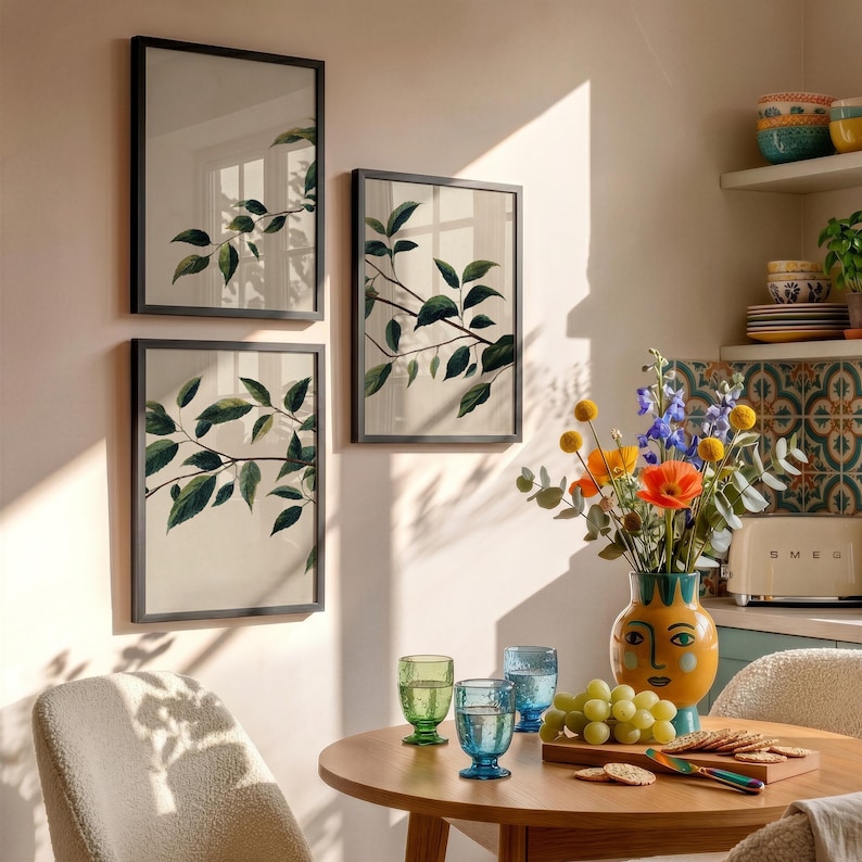 May include: Three framed wall art prints featuring green leaf branches on a light background, displayed above a wooden dining table with a yellow face-shaped vase containing colorful flowers, green and blue glassware, and a plate of crackers.