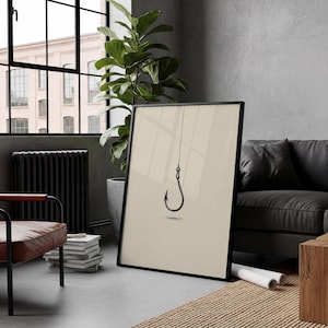 May include: A framed wall art print featuring a minimalist black fishing hook suspended by a thin line against a solid light beige background. The artwork is presented in a thin black frame, showcasing a clean and simple graphic design.