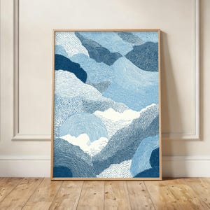 May include: Abstract art print featuring a mountain landscape in shades of blue and white. The artwork is framed in a light wood frame and is displayed against a white wall and wooden floor. The image has a textured, hand-drawn appearance.