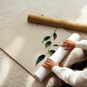 May include: A large, square, off-white poster featuring a single botanical branch with dark green leaves. The poster is partially rolled, with a brown cardboard shipping tube resting on the flat portion of the paper, showcasing a clean, minimalist design.