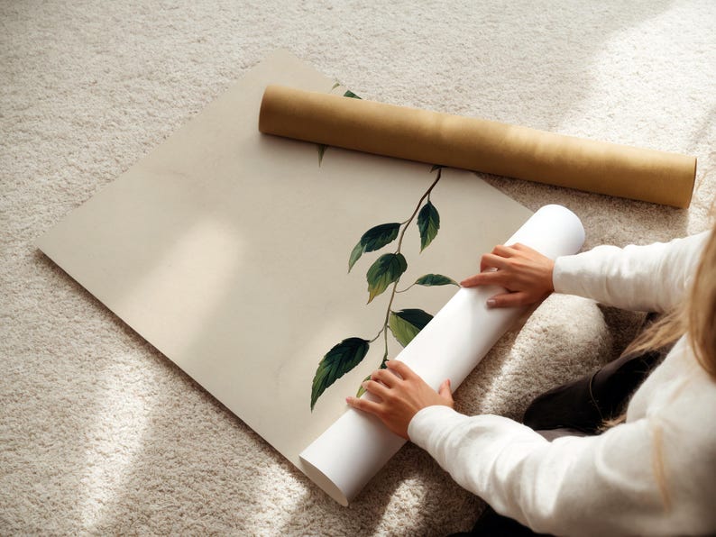 May include: A large, square, off-white poster featuring a single botanical branch with dark green leaves. The poster is partially rolled, with a brown cardboard shipping tube resting on the flat portion of the paper, showcasing a clean, minimalist design.