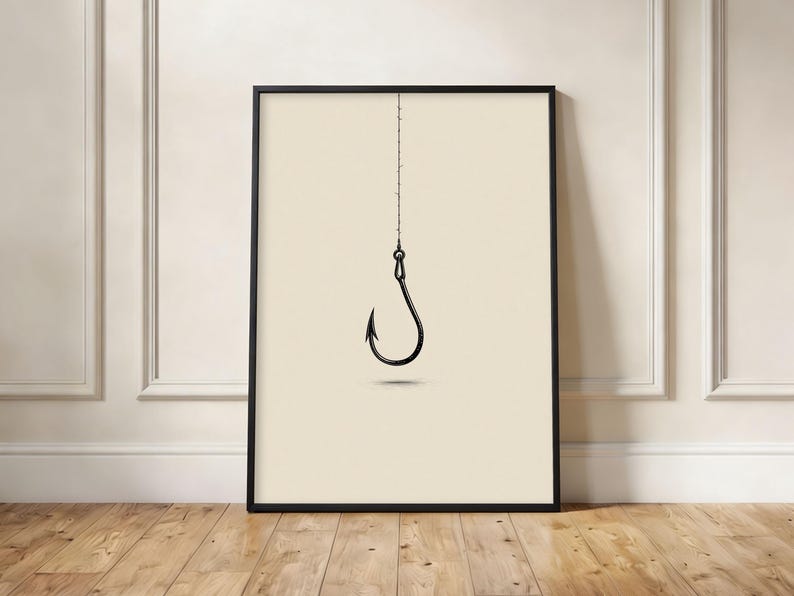 May include: A framed wall art print featuring a black line drawing of a single fishing hook suspended from a thin vertical line. The hook is centered against a solid, light cream-colored background, creating a stark contrast in the composition.