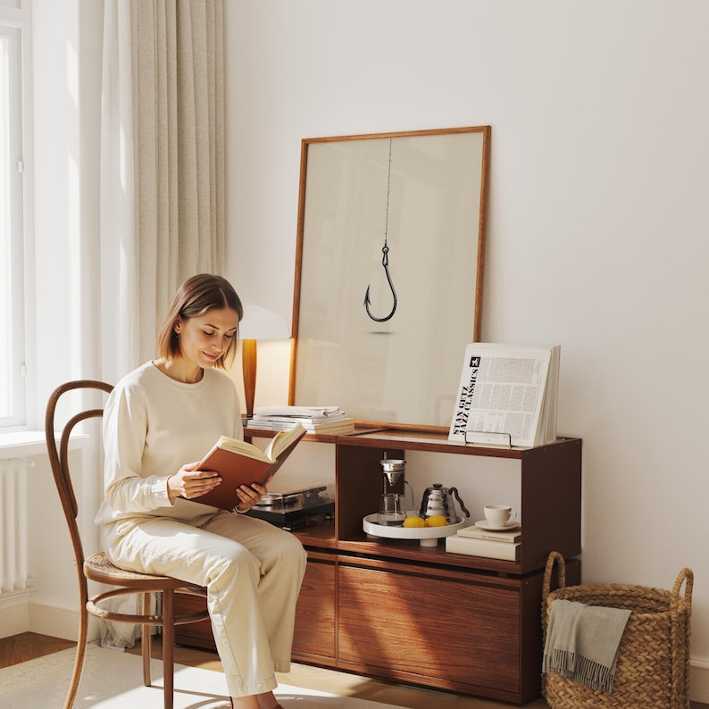 May include: A framed wall art print featuring a black fishing hook illustration on a beige background. The artwork is displayed in a thin wooden frame, positioned on a dark brown wooden cabinet alongside a book, a coffee dripper, and a kettle.