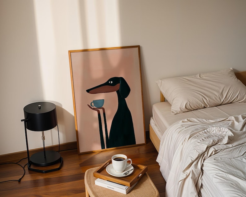 May include: A framed wall art print featuring a stylized illustration of a black and tan dog with long ears holding a small light blue cup. The dog is set against a solid light pink background, and the artwork is enclosed in a thin wooden frame.