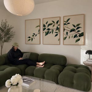 May include: A modular olive green tufted sofa with a low profile and rounded segments. Three framed vertical wall art prints feature green leafy branches on a cream background. A clear glass coffee table holds a white vase with white flowers.