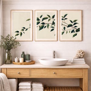 May include: A set of three framed wall art prints featuring botanical illustrations of green leafy branches against a cream background, displayed above a bathroom vanity with a white vessel sink, a glass vase of greenery, and stacked towels.