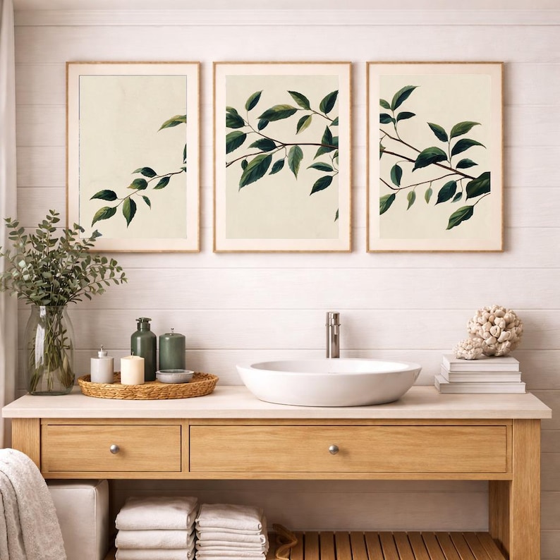May include: A set of three framed wall art prints featuring botanical illustrations of green leafy branches against a cream background, displayed above a bathroom vanity with a white vessel sink, a glass vase of greenery, and stacked towels.