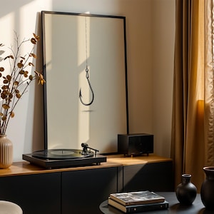 May include: A framed vertical wall art print featuring a black line drawing of a single fishing hook hanging from a thin vertical line. The artwork is displayed on a wooden cabinet next to a black record player, a small speaker, and a ribbed vase.