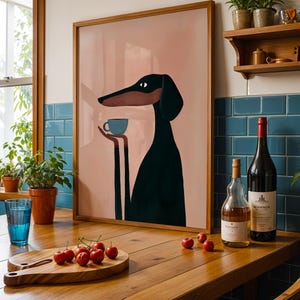 May include: A framed wall art print featuring a stylized black and brown dog with long ears holding a small light blue cup. The illustration is set against a solid dusty pink background, presented in a simple wooden frame on a kitchen counter.