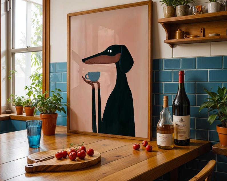 May include: A framed wall art print featuring a stylized black and brown dog with long ears holding a small light blue cup. The illustration is set against a solid dusty pink background, presented in a simple wooden frame on a kitchen counter.
