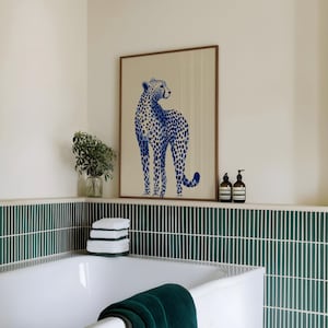 May include: A framed wall art print featuring a blue cheetah illustration with a spotted pattern on a light beige background. The artwork is displayed in a wooden frame, positioned above a bathtub with dark green vertical tiled walls.