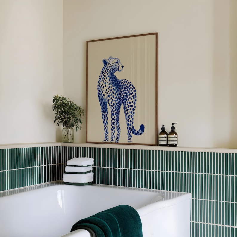 May include: A framed wall art print featuring a blue cheetah illustration with a spotted pattern on a light beige background. The artwork is displayed in a wooden frame, positioned above a bathtub with dark green vertical tiled walls.