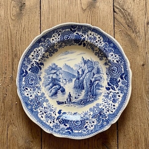 Peut inclure: Une assiette ronde antique bleue et blanche avec un bord festonné. L'assiette présente une scène de paysage détaillée avec un château, des personnages dans un bateau et une bordure florale. L'assiette est posée sur une surface en bois.
